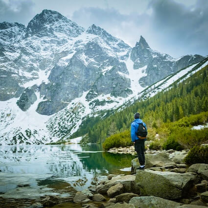 Explorador parado al borde de un lago frente a una montaña con nieve